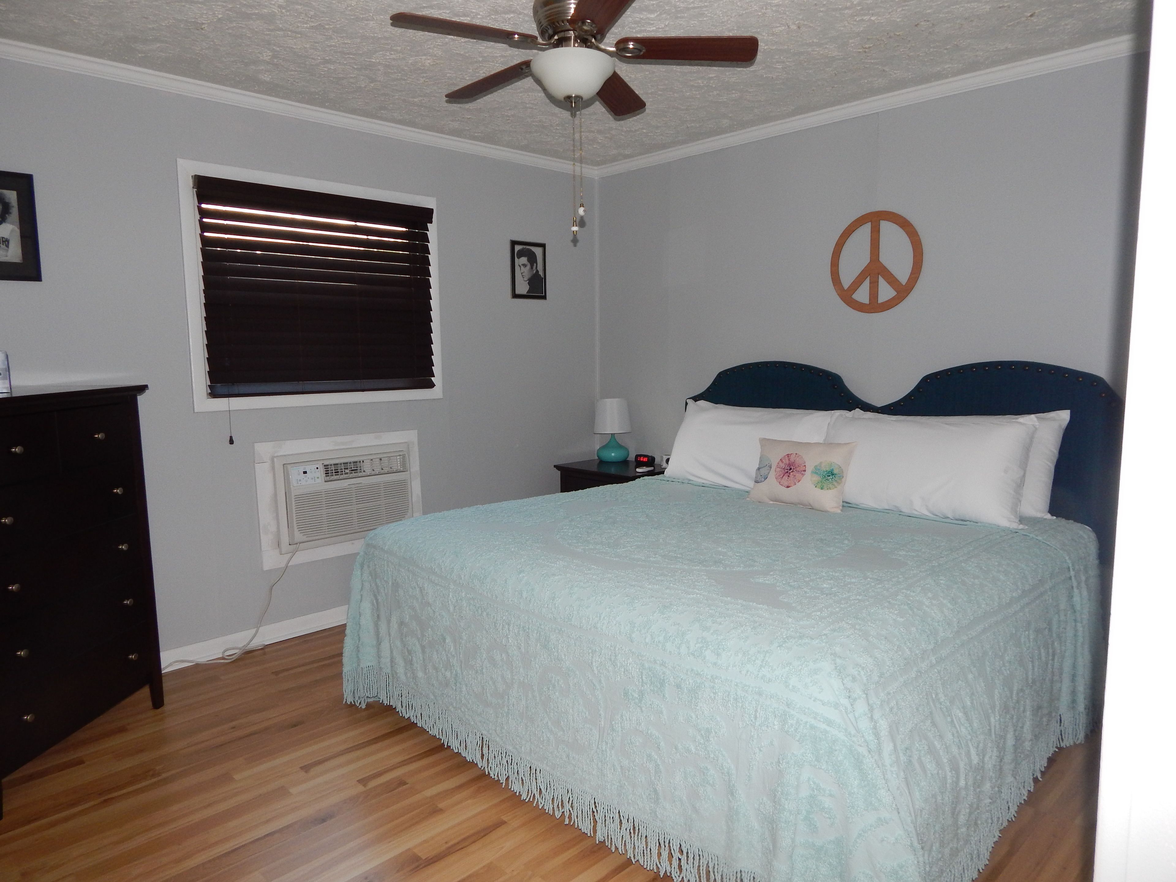 Un dormitorio con cama, ventilador de techo, ventana con persianas, aire acondicionado de pared, mesita de noche con lámpara y un símbolo de la paz en la pared.