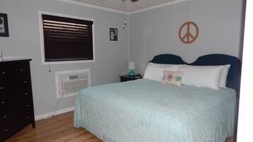 Un dormitorio con cama, ventilador de techo, ventana con persianas, aire acondicionado de pared, mesita de noche con lámpara y un símbolo de la paz en la pared.