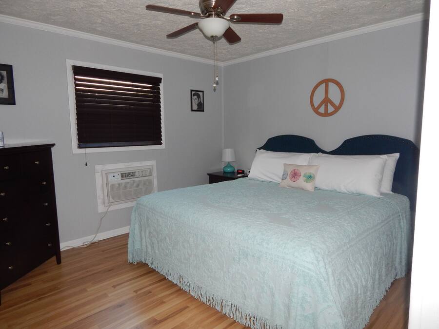Un dormitorio con cama, ventilador de techo, ventana con persianas, aire acondicionado de pared, mesita de noche con lámpara y un símbolo de la paz en la pared.