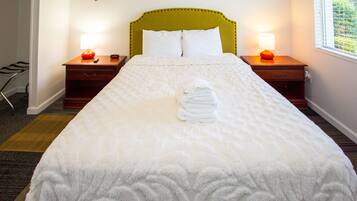 A neatly made bed with white bedding, two bedside tables with lamps, a wooden headboard, and a peace sign decoration on the wall.