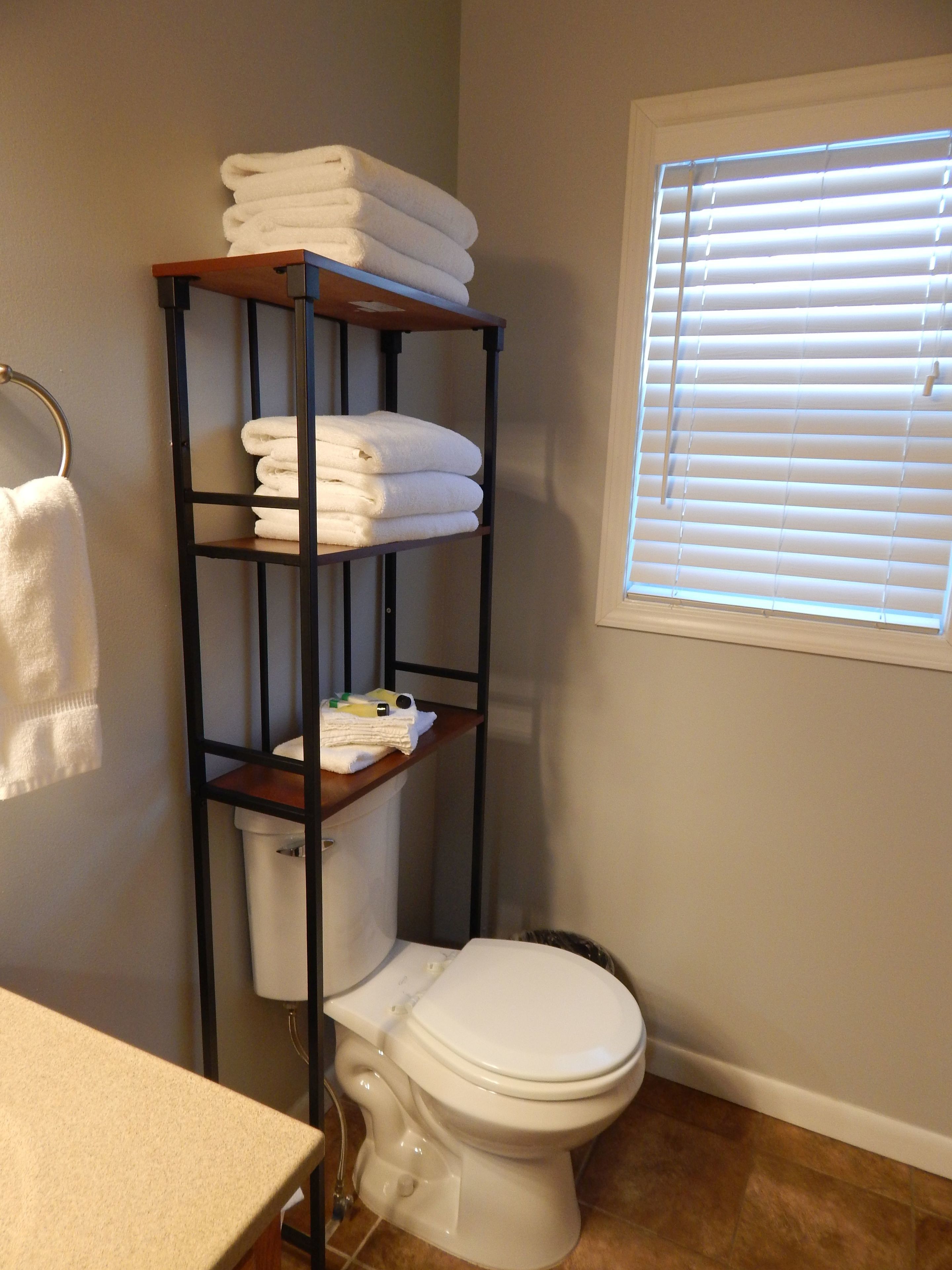A bathroom with a toilet, a shelf with folded towels, and a window with blinds.