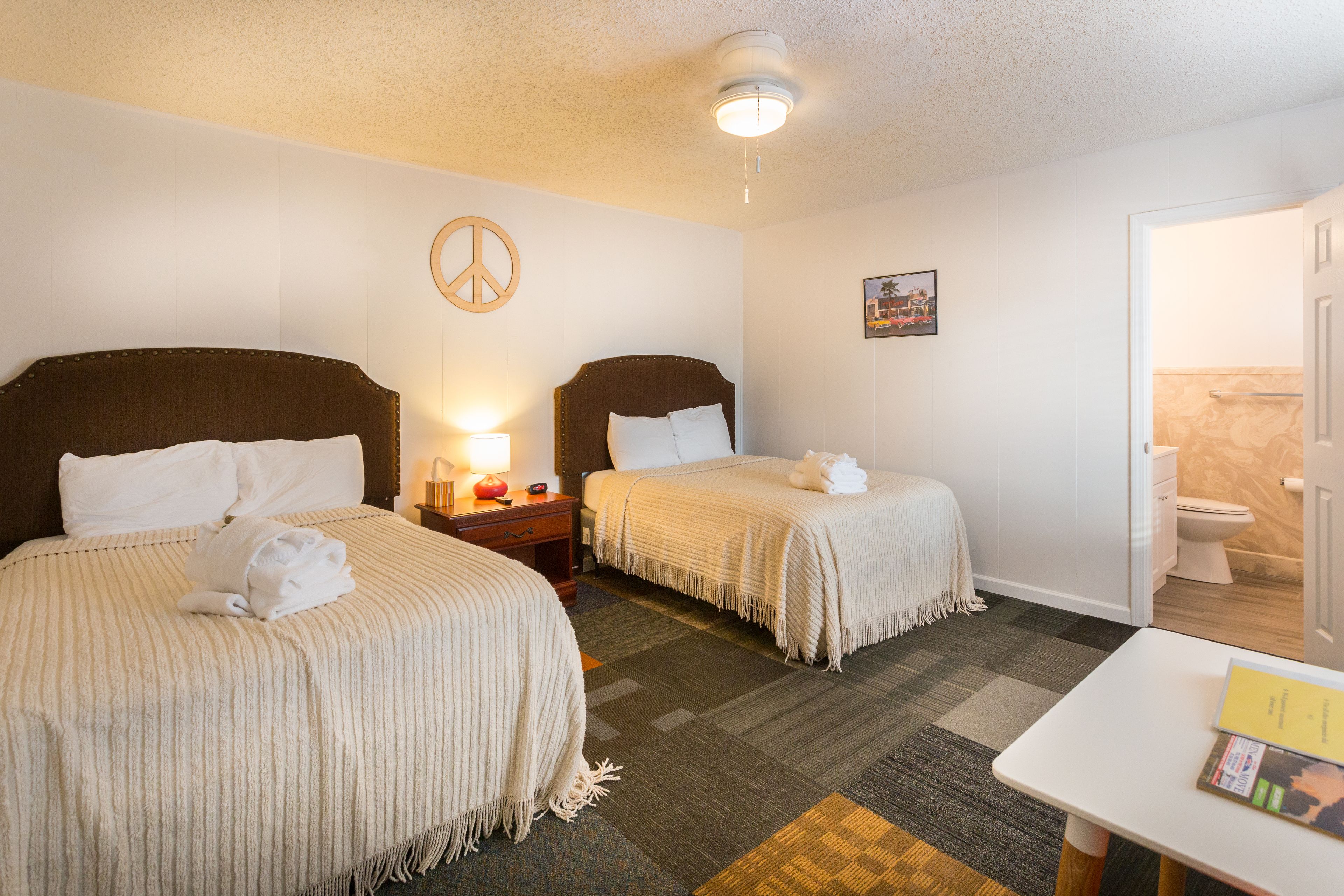 A hotel room with two beds, a small table, a peace sign on the wall, and a bathroom visible through an open door.