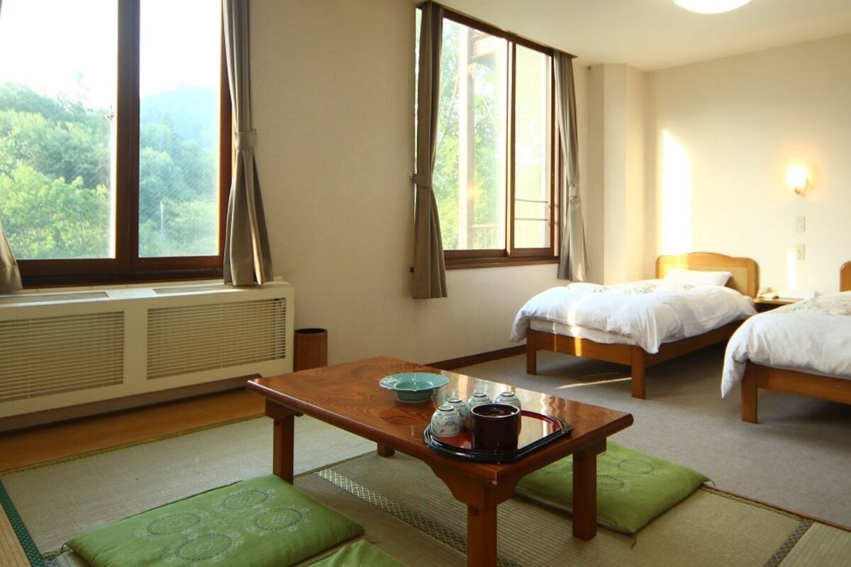 Une chambre d’hôtel avec deux lits, une table en bois avec un plateau, un radiateur et une vue sur la verdure à l’extérieur.