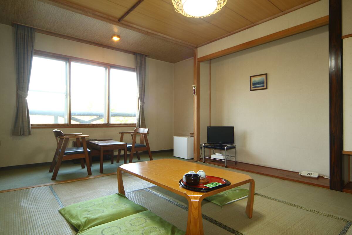 Une pièce de style japonais traditionnel, avec un sol recouvert de tatamis, une petite table en bois, un téléviseur et une fenêtre avec des rideaux.