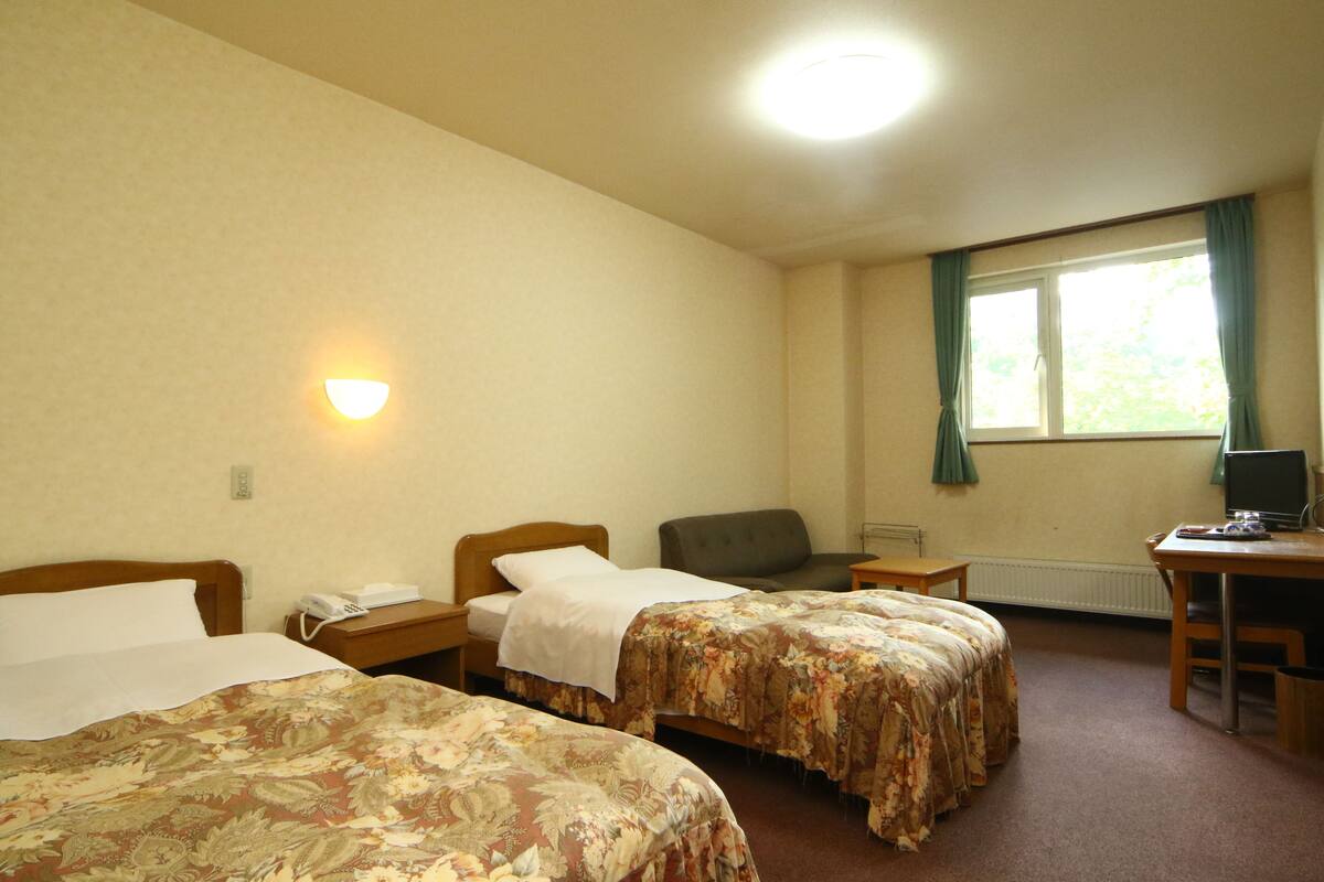 Une chambre d’hôtel avec deux lits, un bureau, une chaise, un ordinateur, un téléphone et une fenêtre avec des rideaux.