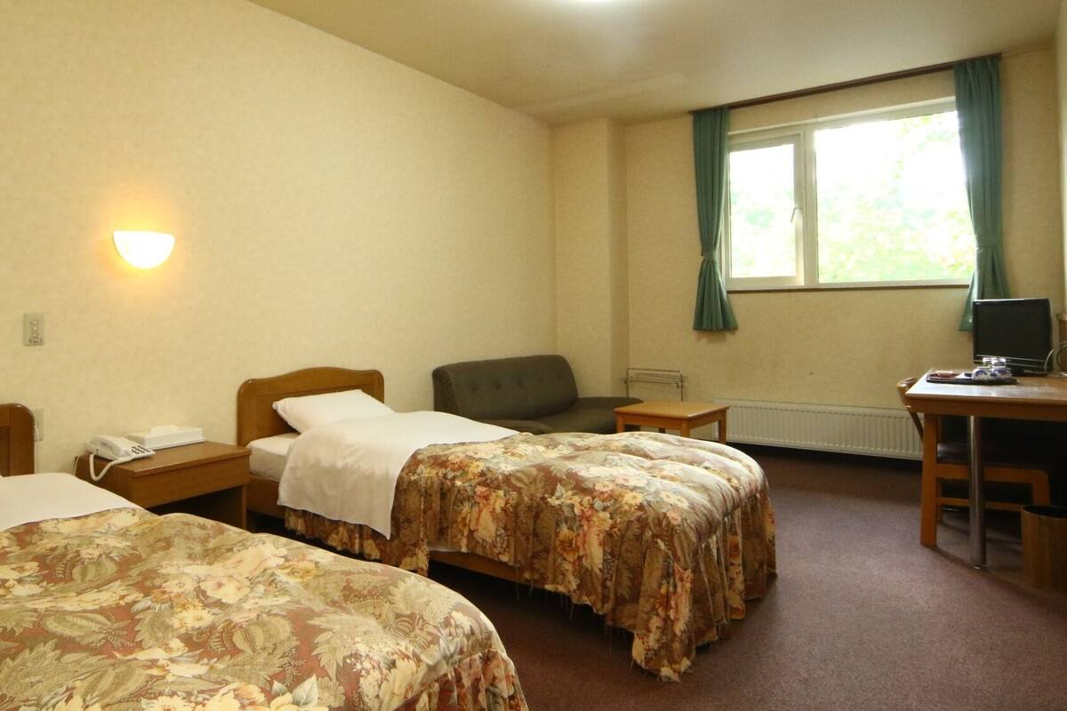 Une chambre d’hôtel avec deux lits, un bureau avec un ordinateur, une chaise, un téléphone et une fenêtre avec des rideaux.