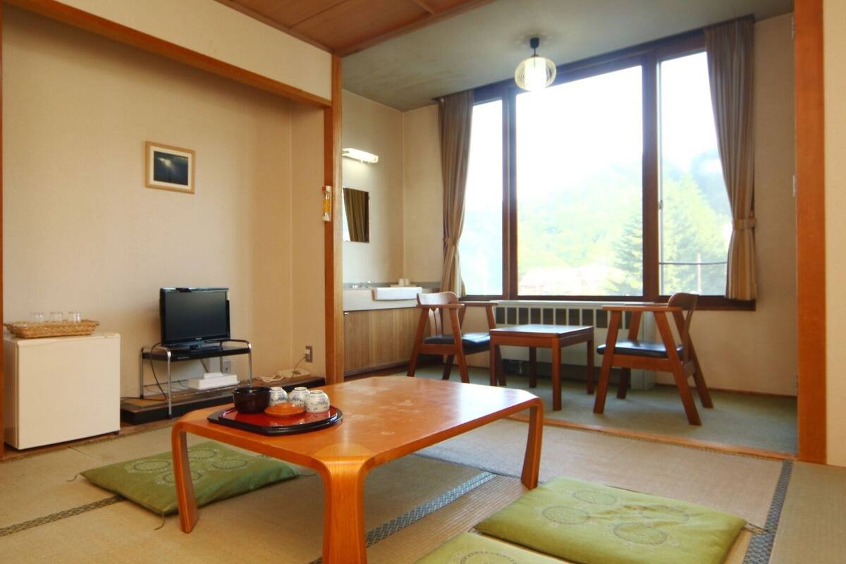 Une pièce comprenant un téléviseur posé sur un meuble, une petite armoire blanche, une table en bois avec un plateau, et une fenêtre donnant sur des arbres.