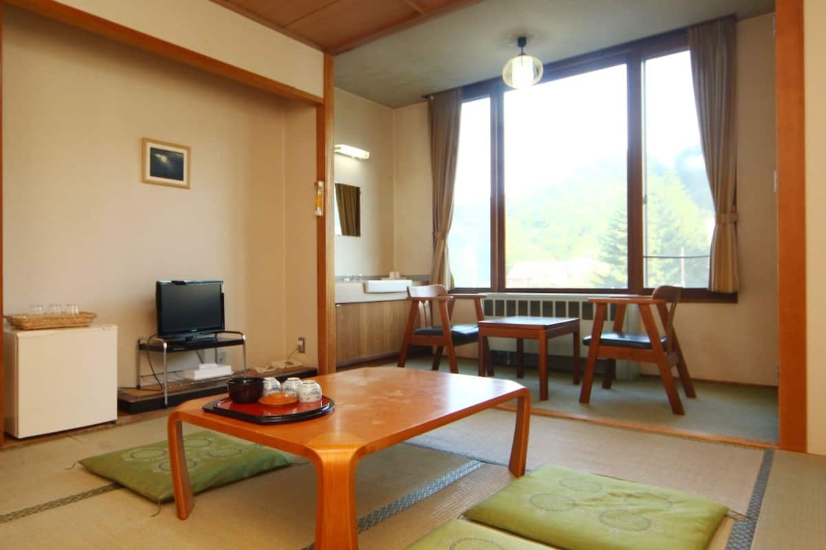 Une pièce comprenant un téléviseur posé sur un meuble, une petite armoire blanche, une table en bois avec un plateau, et une fenêtre donnant sur des arbres.