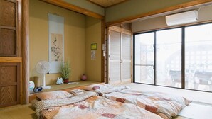 A traditional Japanese room with tatami flooring, a futon bed, sliding shoji doors, a fan, and a view through the shoji doors. - ALL FRIENDS - Hostel (Nago)