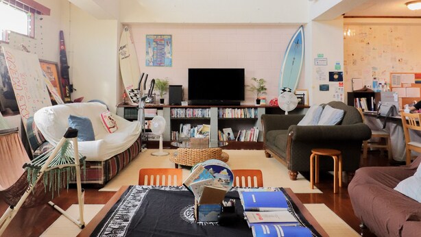 A spacious living room with a large flat-screen TV, a surfboard, and a bookshelf filled with books. - ALL FRIENDS - Hostel (Nago)
