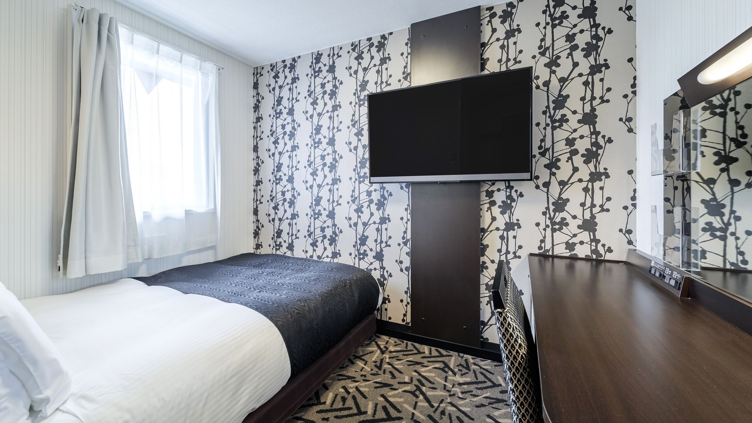 A hotel room with a bed, a TV mounted on the wall, a wooden dresser, and a patterned carpet.