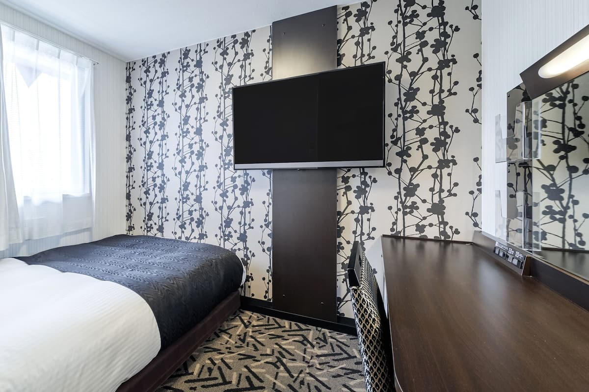 A hotel room with a bed, a TV mounted on the wall, a wooden dresser, and a patterned carpet.