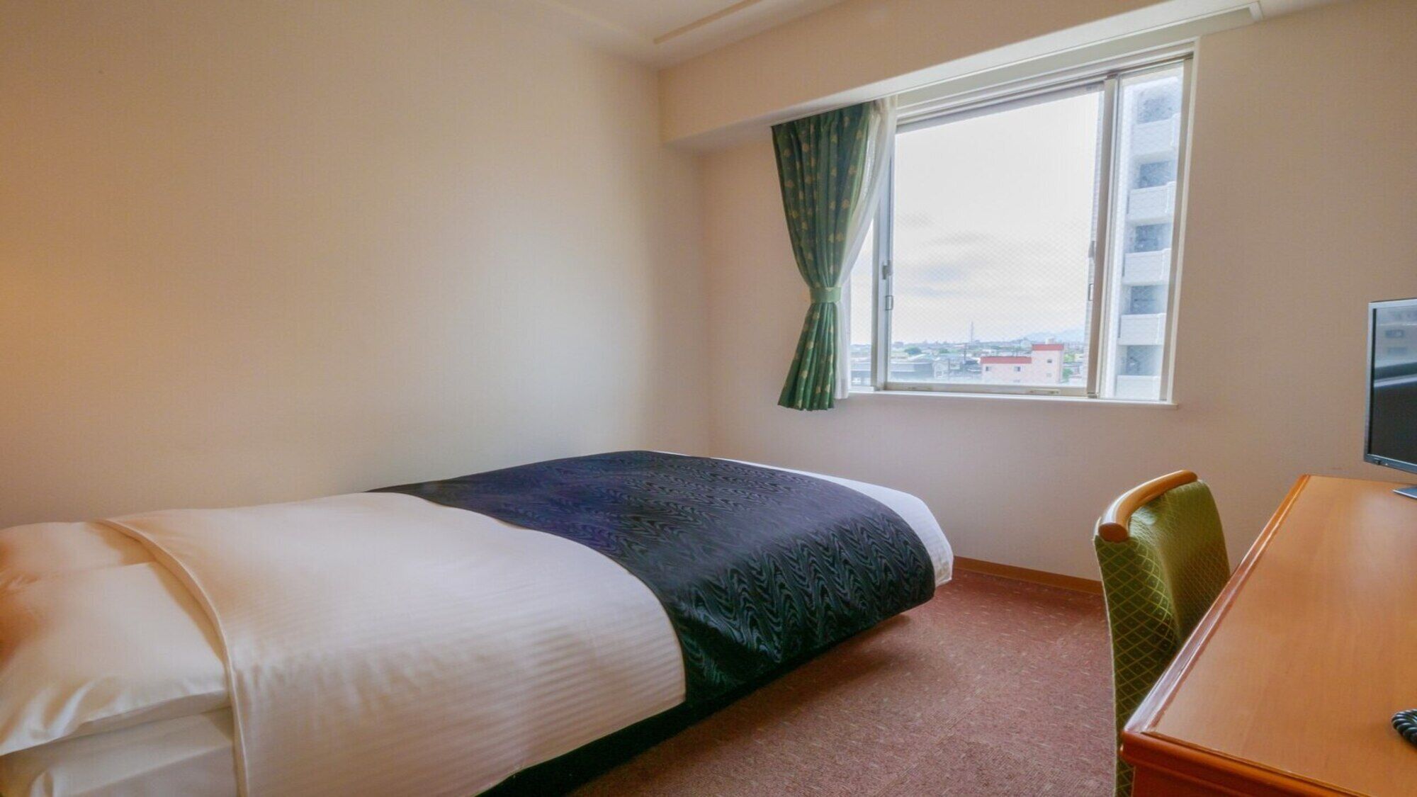 A hotel room with a bed, a desk, a chair, a window with curtains, and a view of buildings outside.