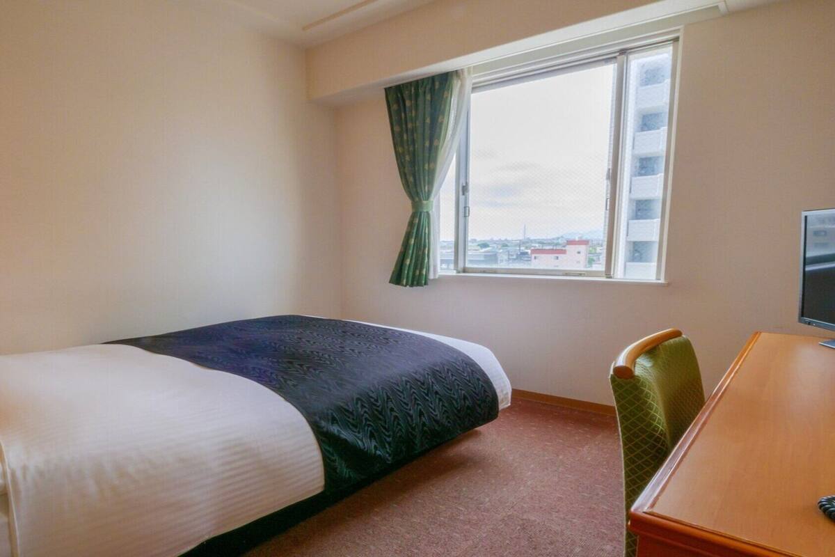 A hotel room with a bed, a desk, a chair, a window with curtains, and a view of buildings outside.