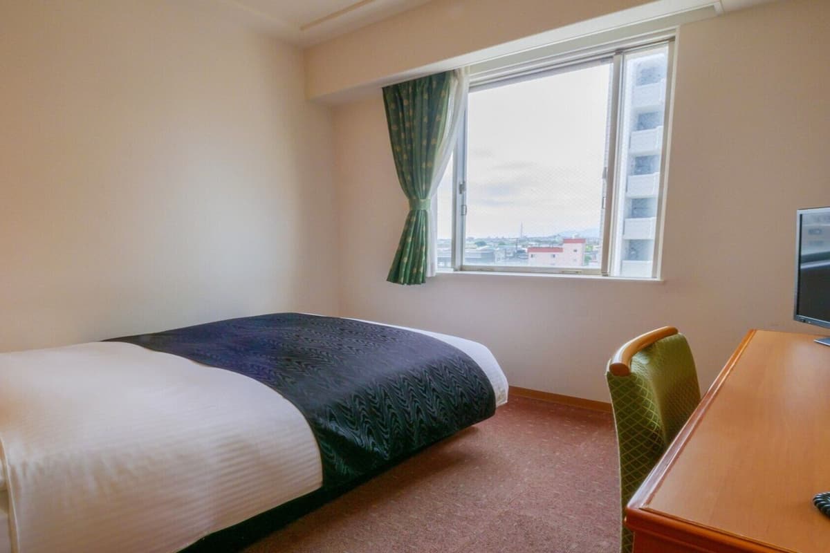 A hotel room with a bed, a desk, a chair, a window with curtains, and a view of buildings outside.