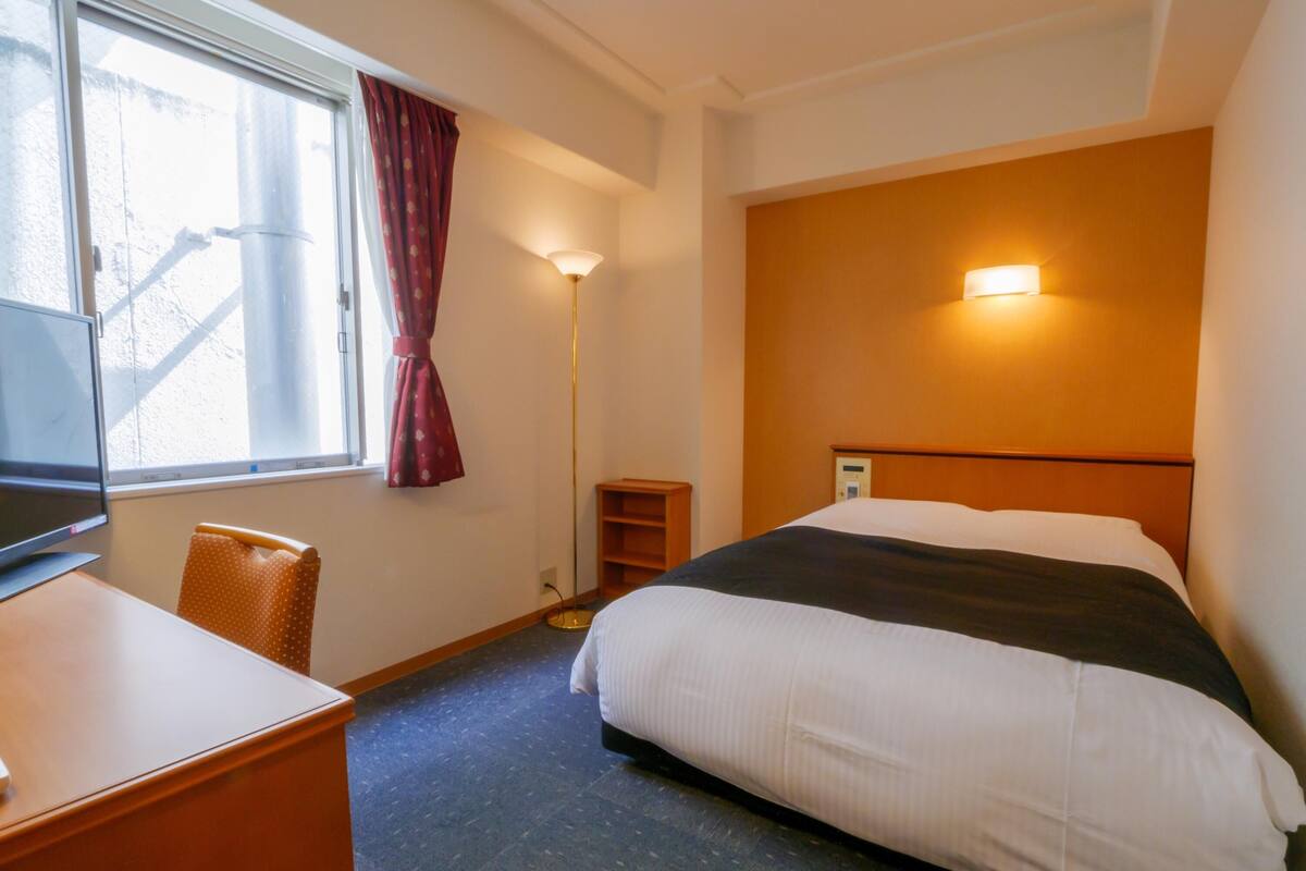 A hotel room with a bed, a desk, a chair, a TV, a window with curtains, and a wall-mounted light.