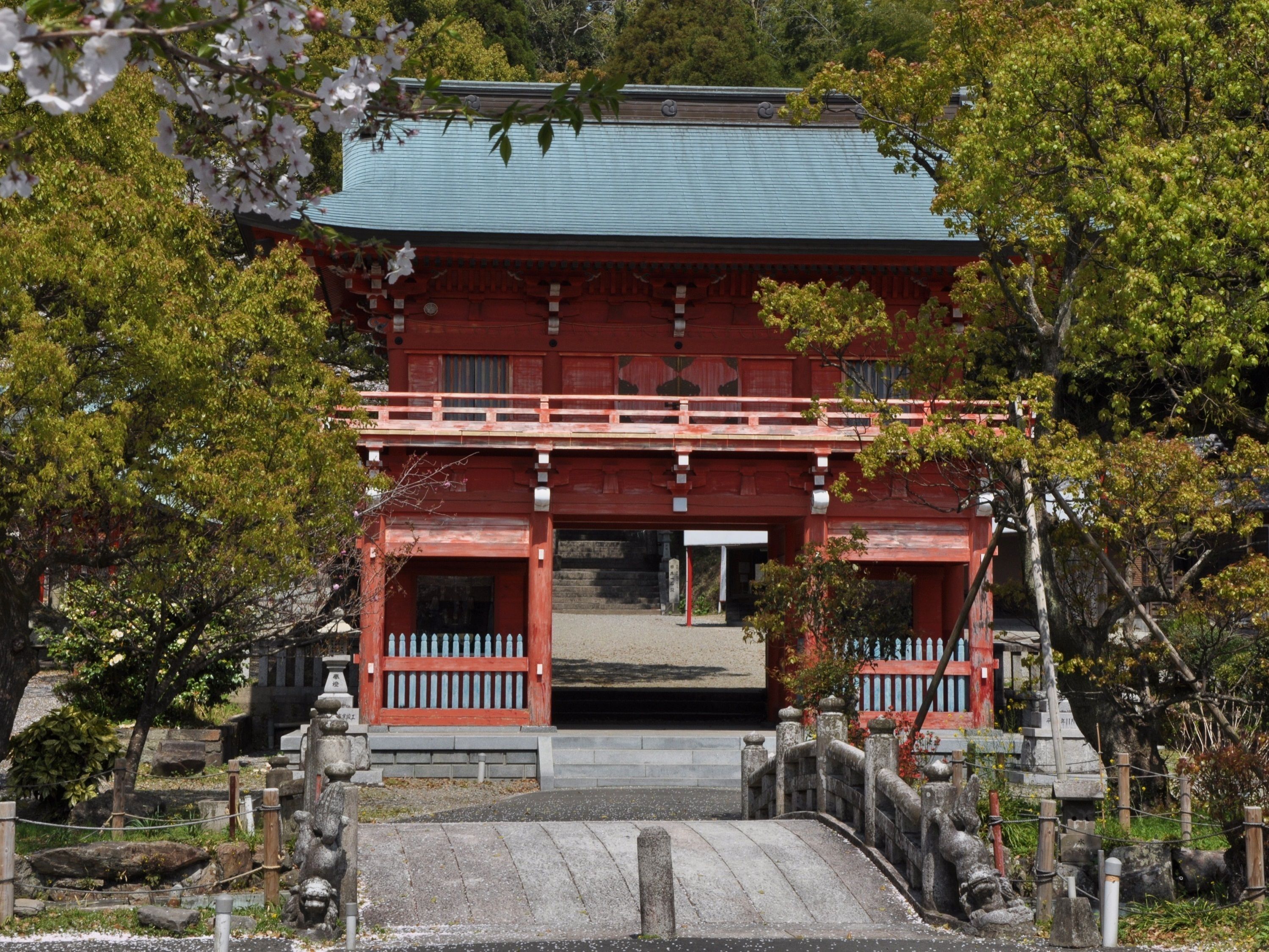 Un bâtiment traditionnel japonais, avec une façade rouge et un toit vert, entouré d’arbres et surplombant un pont de pierre.