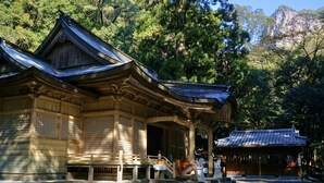 A traditional Japanese building with a tiled roof and wooden structure, surrounded by trees and a mountain in the background. - APA Hotel Miyazaki Nobeoka Chuo (Nobeoka)