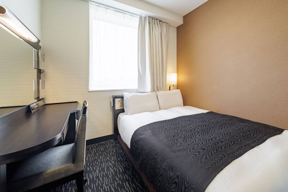 A hotel room with a bed, a desk, a chair, a lamp, and a window with curtains.