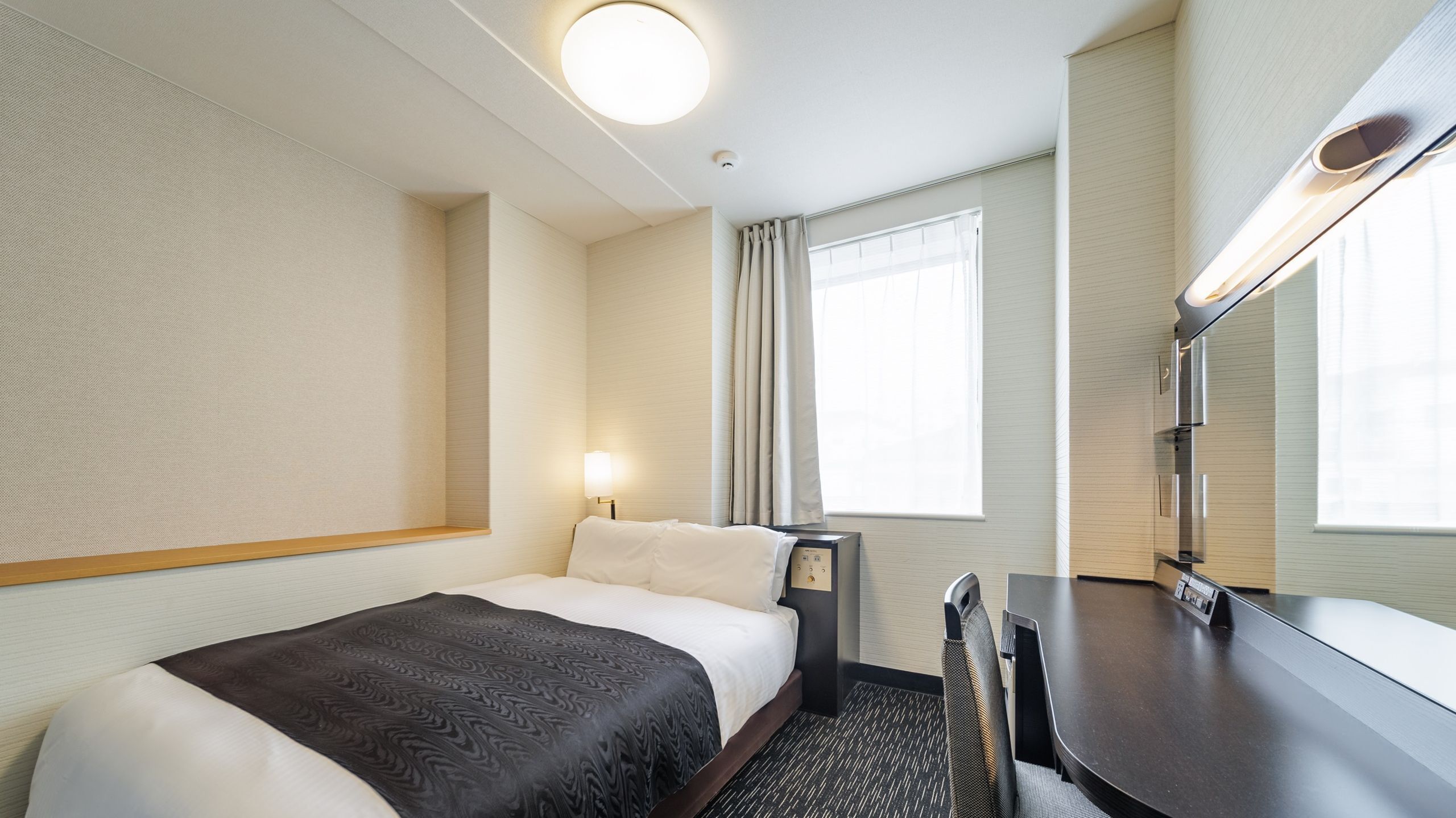 A hotel room with a bed, bedside table, lamp, desk, chair, and a window with curtains.
