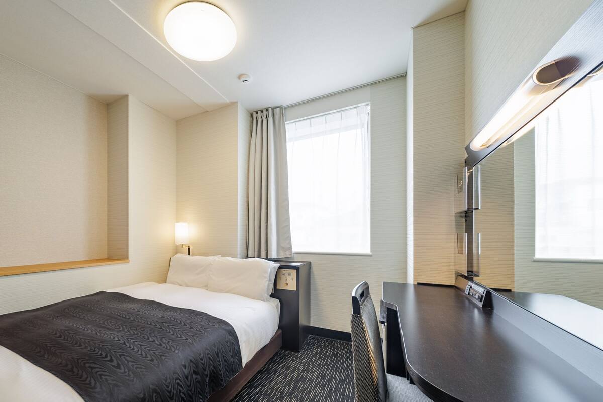 A hotel room with a bed, bedside table, lamp, desk, chair, and a window with curtains.