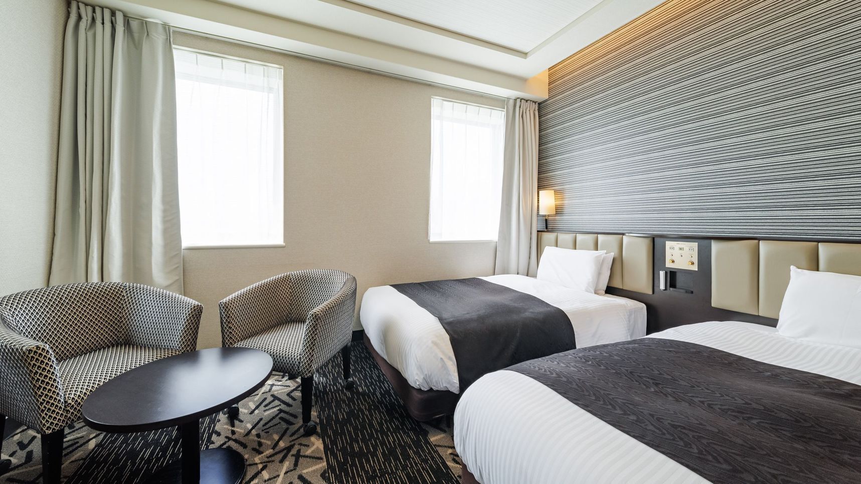 A hotel room with two beds, a sofa, a small table, and a wall-mounted lamp.