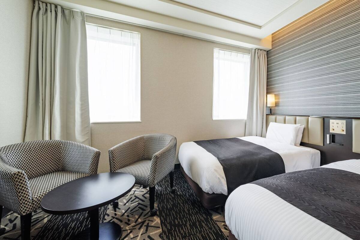 A hotel room with two beds, a sofa, a small table, and a wall-mounted lamp.