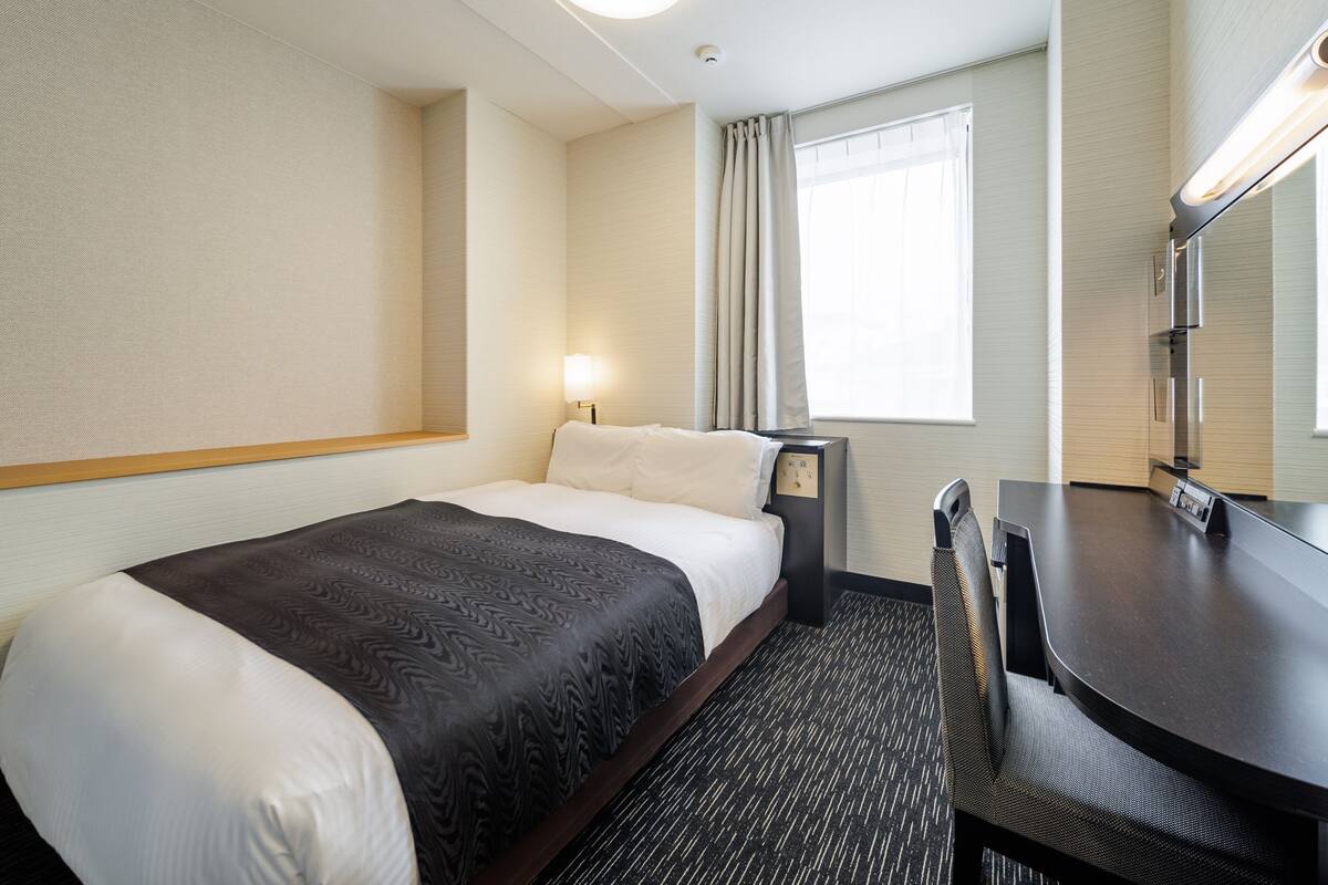 A hotel room with a bed, a desk, a chair, a desk lamp, a window with curtains, and a wall-mounted light fixture.