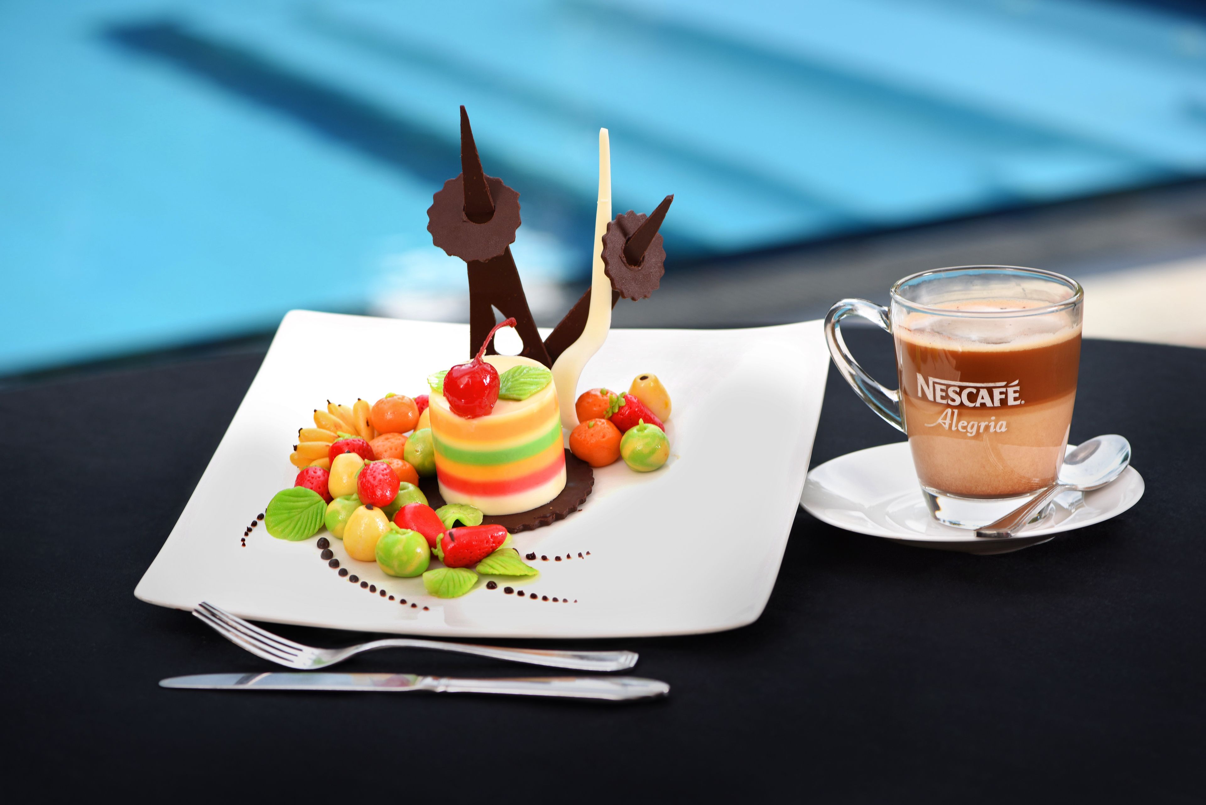 A dessert plate with colorful fruit and chocolate decorations, a glass of Nescafé coffee, and cutlery on a table.