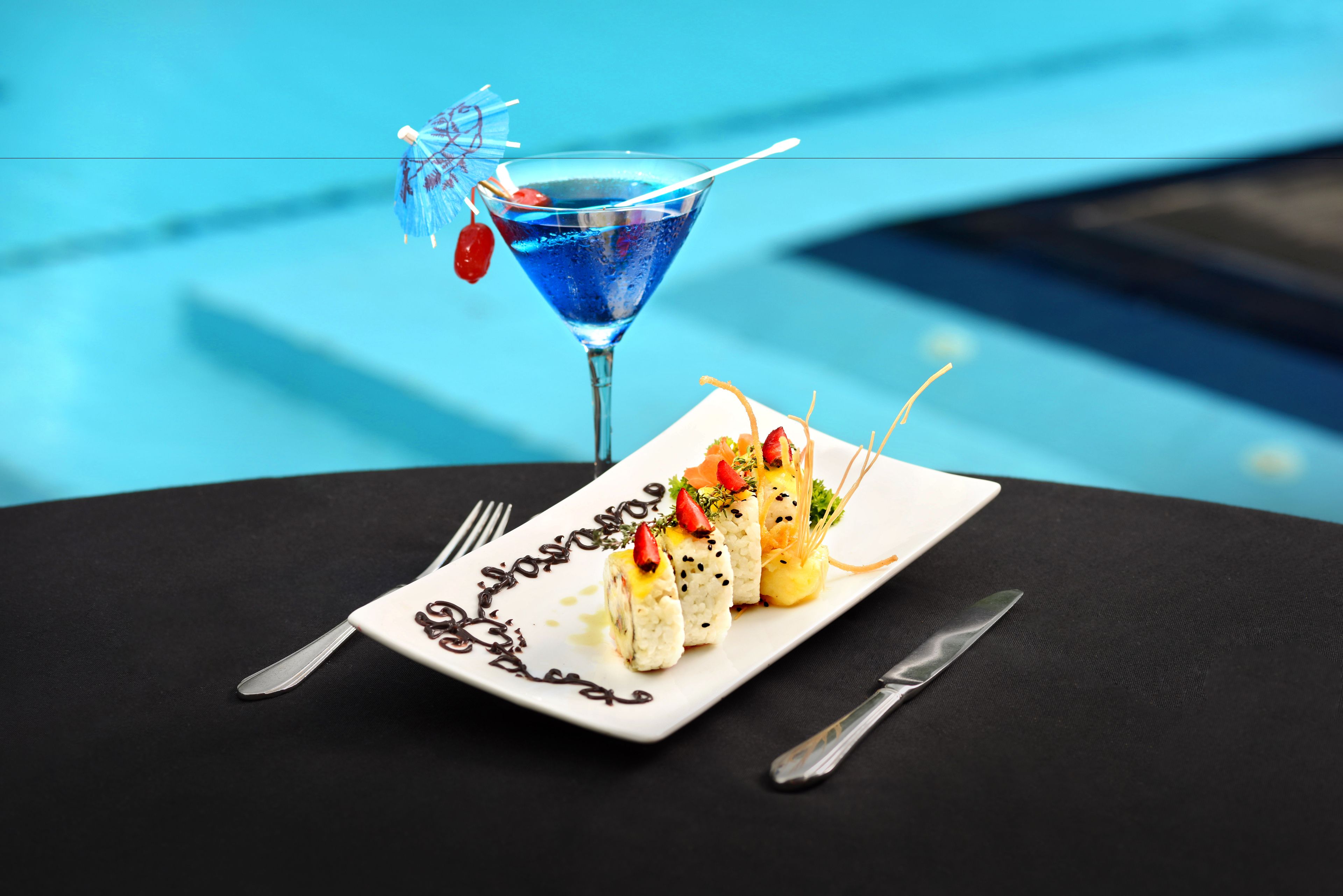 A dining table with a white plate of sushi, a blue cocktail with a straw, and a fork and knife on the table.