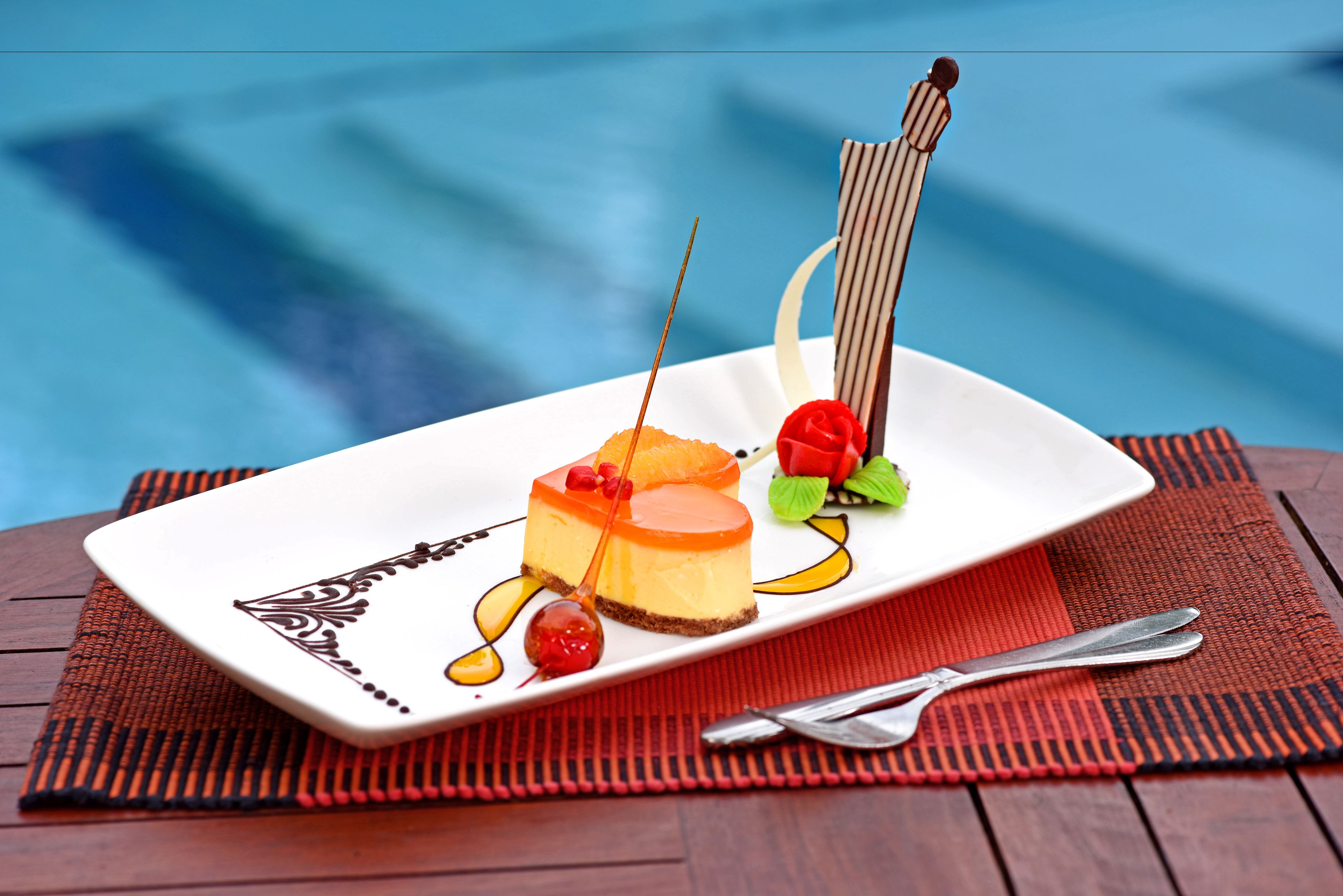 A dessert on a white plate with a striped napkin and a spoon on a wooden table by a pool.
