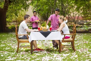 A group of people dining outdoors at a table with a white tablecloth, surrounded by greenery and a pond.
