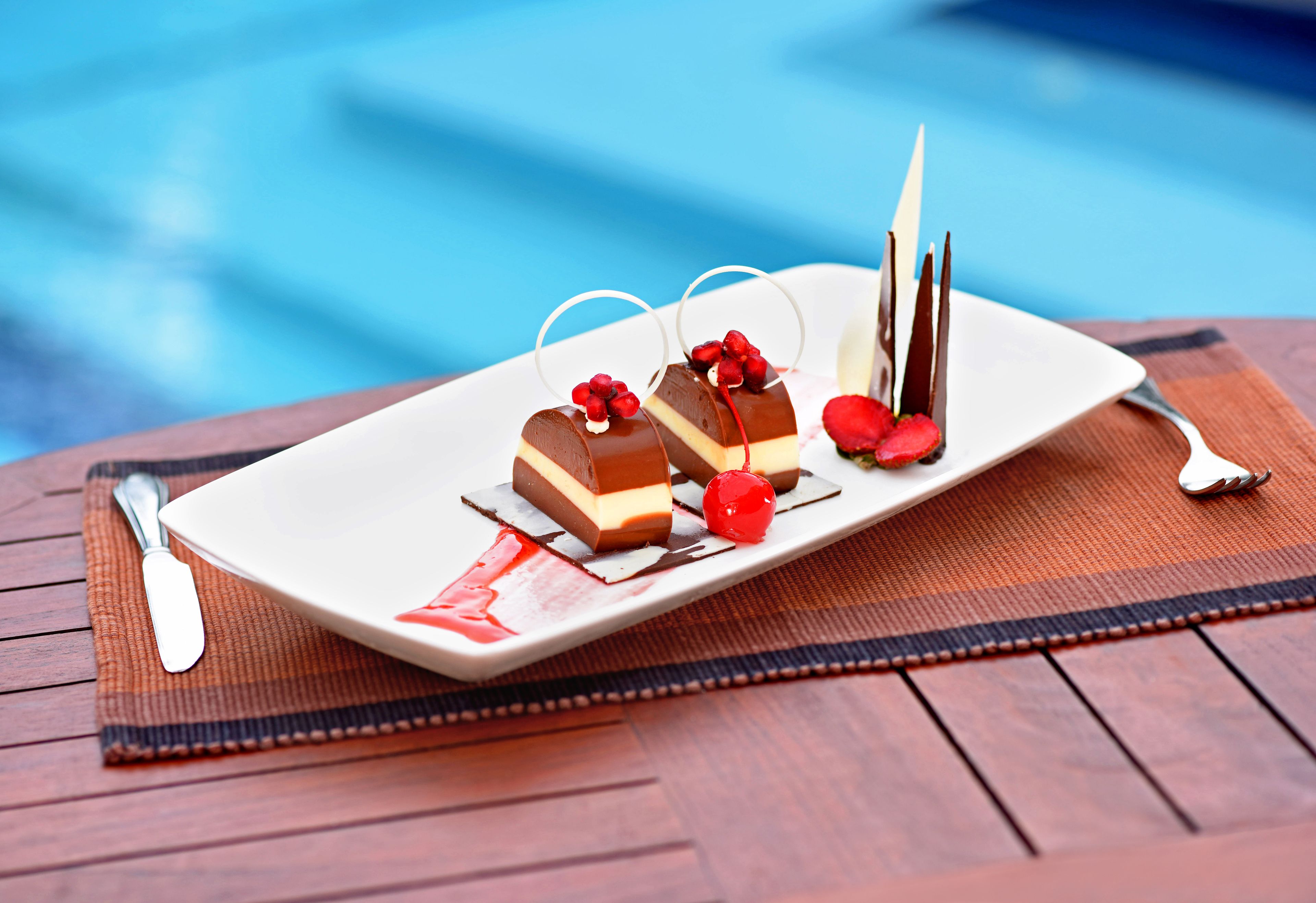 Eine Dessertplatte mit Schokoladen- und Cremeschichten, Kirschen und einer Schokoladendekoration auf einem Holztisch in der Nähe eines Pools.