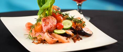 A square white plate with a seafood dish, a cocktail with a lemon garnish, and a dark tablecloth against a blue background.