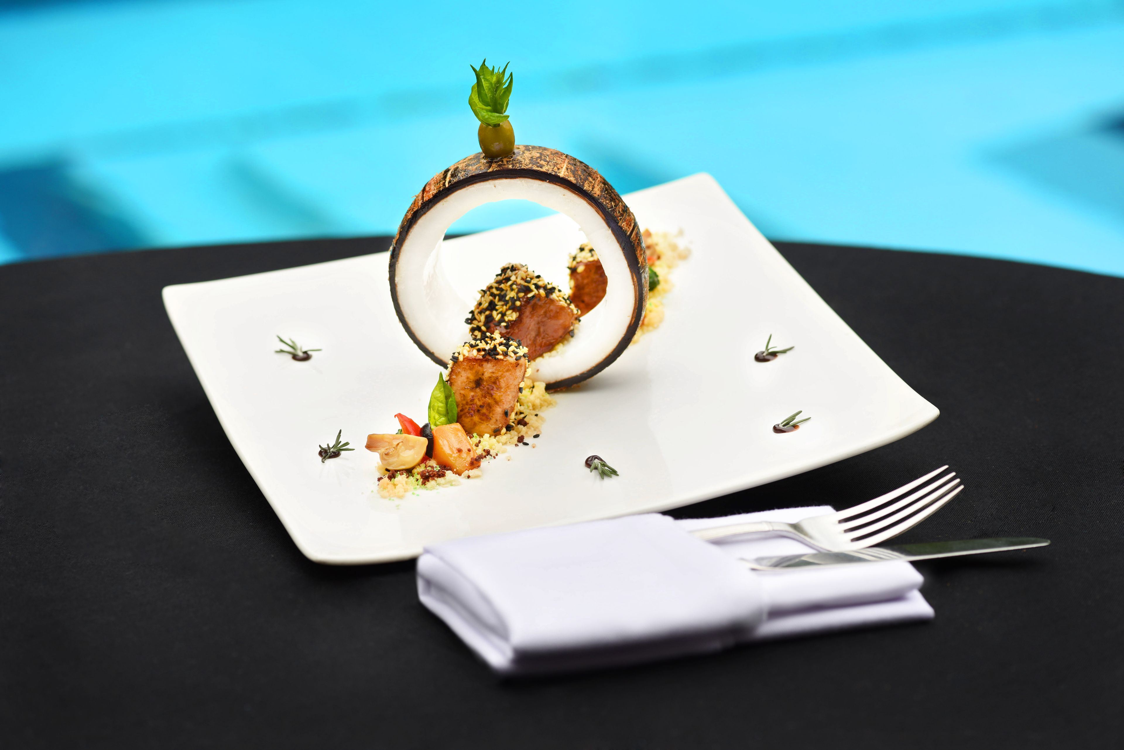 Un plato blanco con una presentación original de comida, que incluye carne, verduras y un adorno que recuerda a una cáscara de coco.