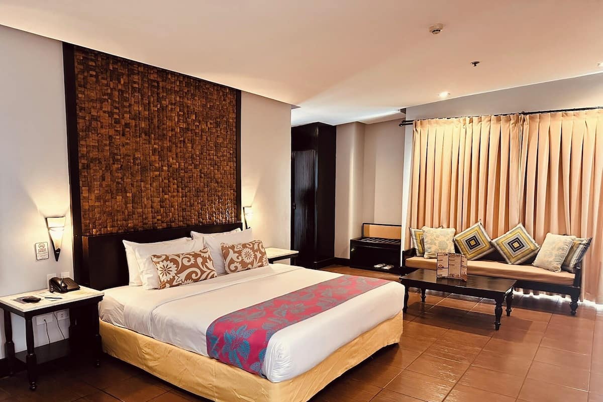 A hotel room with a large bed, a bedside table with a lamp, a sofa with cushions, and a curtain-covered window.