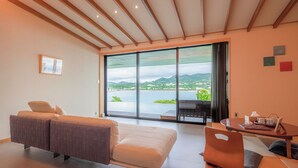 A modern room with a sofa, a table with a teapot, and a view of a lake and mountains through large windows. - Sesoko Sansui (Motobu)