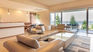 A modern living room with a sofa set, a coffee table, and a dining area with a table and chairs. There is a balcony with a railing and greenery visible through the windows. - Condominium T-Room (Yomitan)