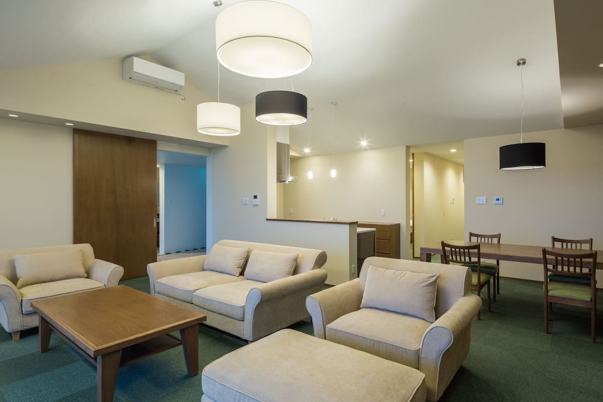 A modern hotel room with a sofa set, a wooden coffee table, a dining table with chairs, and pendant lighting.