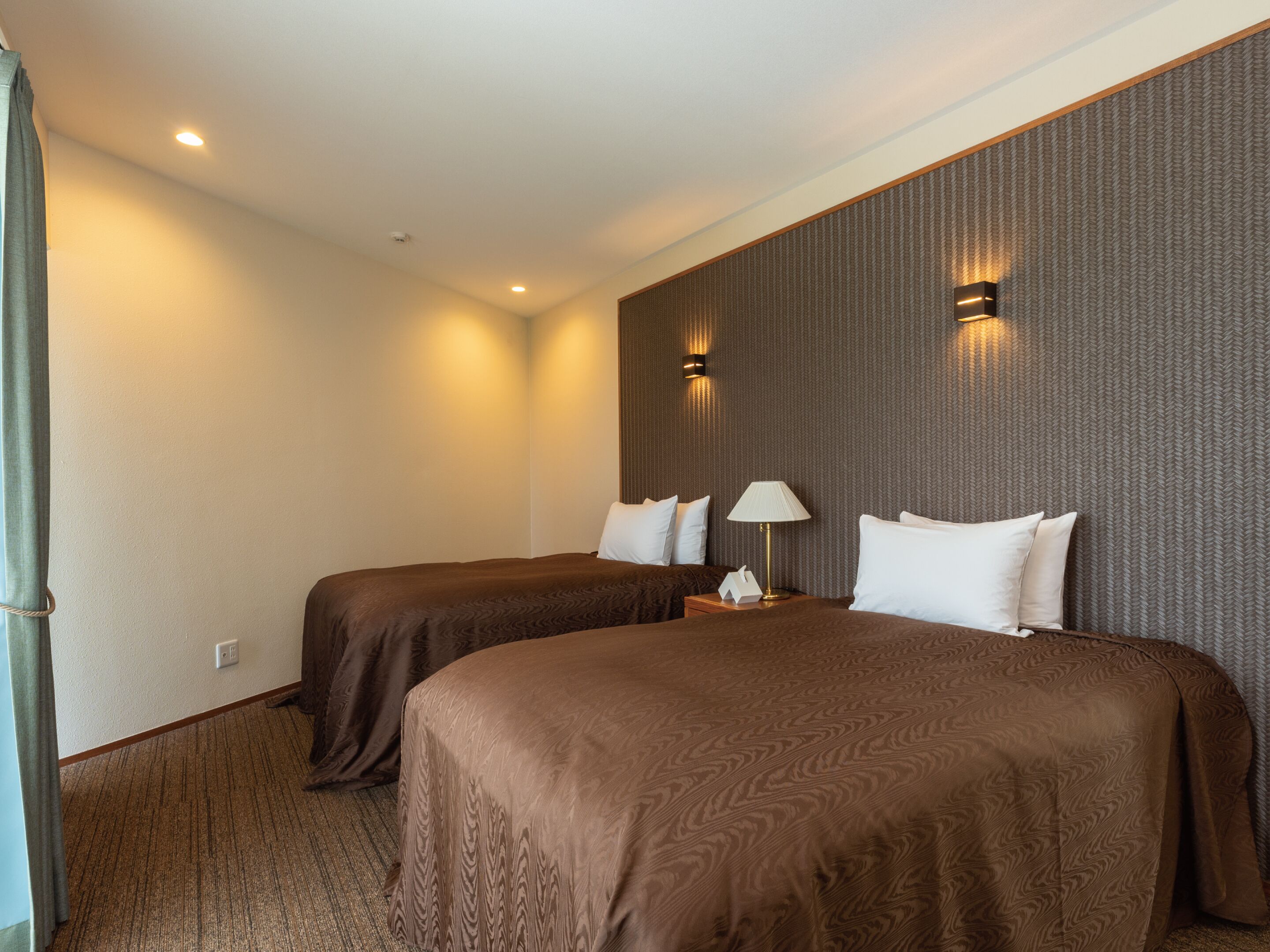 A hotel room with two beds, a nightstand with a lamp, and a wall-mounted light fixture.