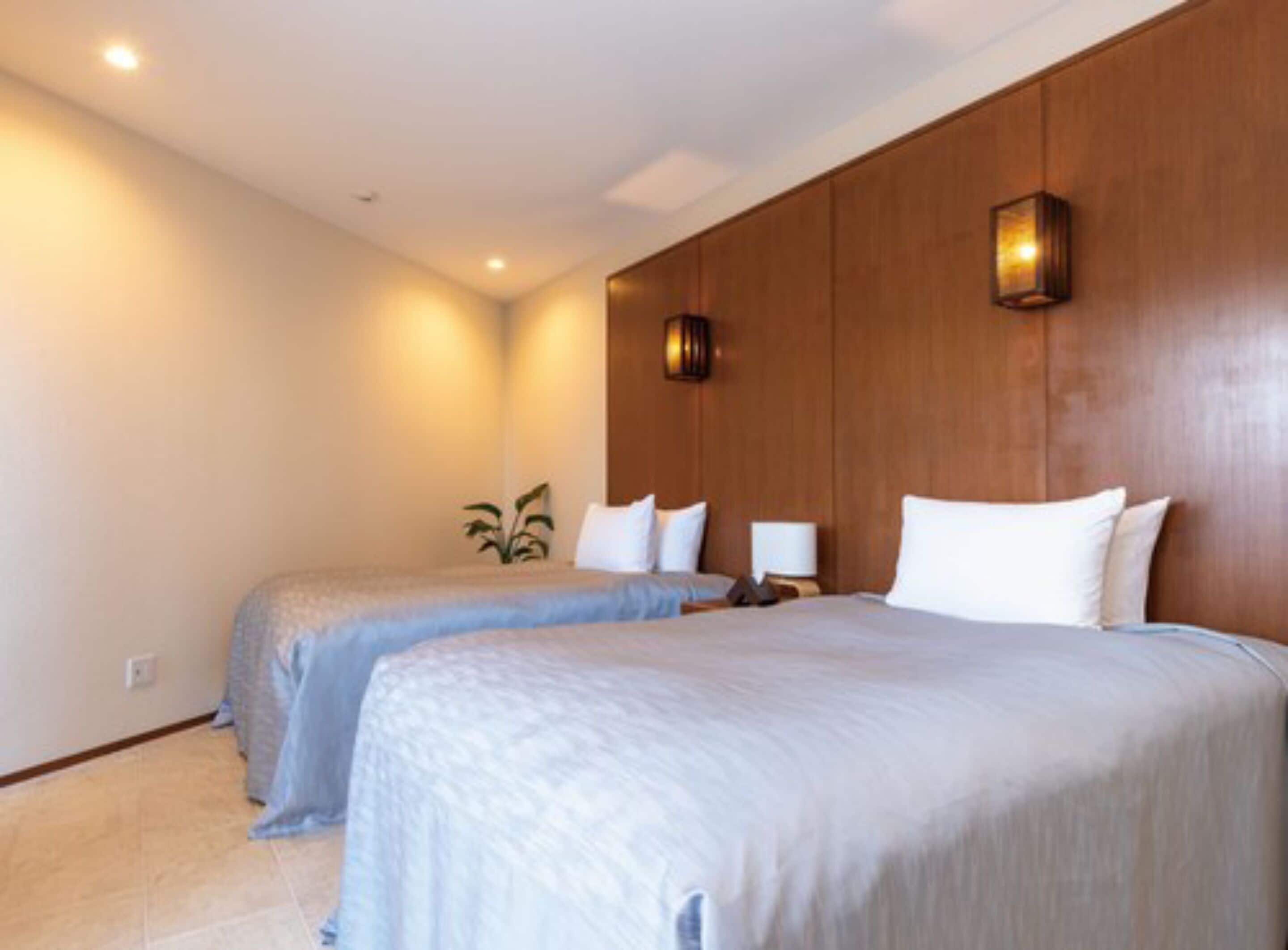 A hotel room with two beds, a wooden headboard, a wall-mounted lamp, and a small plant.