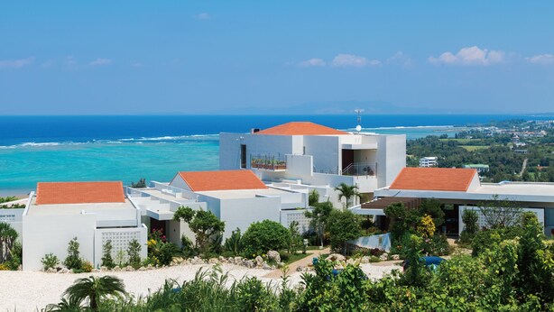 A coastal property with modern architecture, featuring multiple buildings with red-tiled roofs and white walls, surrounded by lush greenery and overlooking the ocean. - Condominium T-Room (Yomitan)