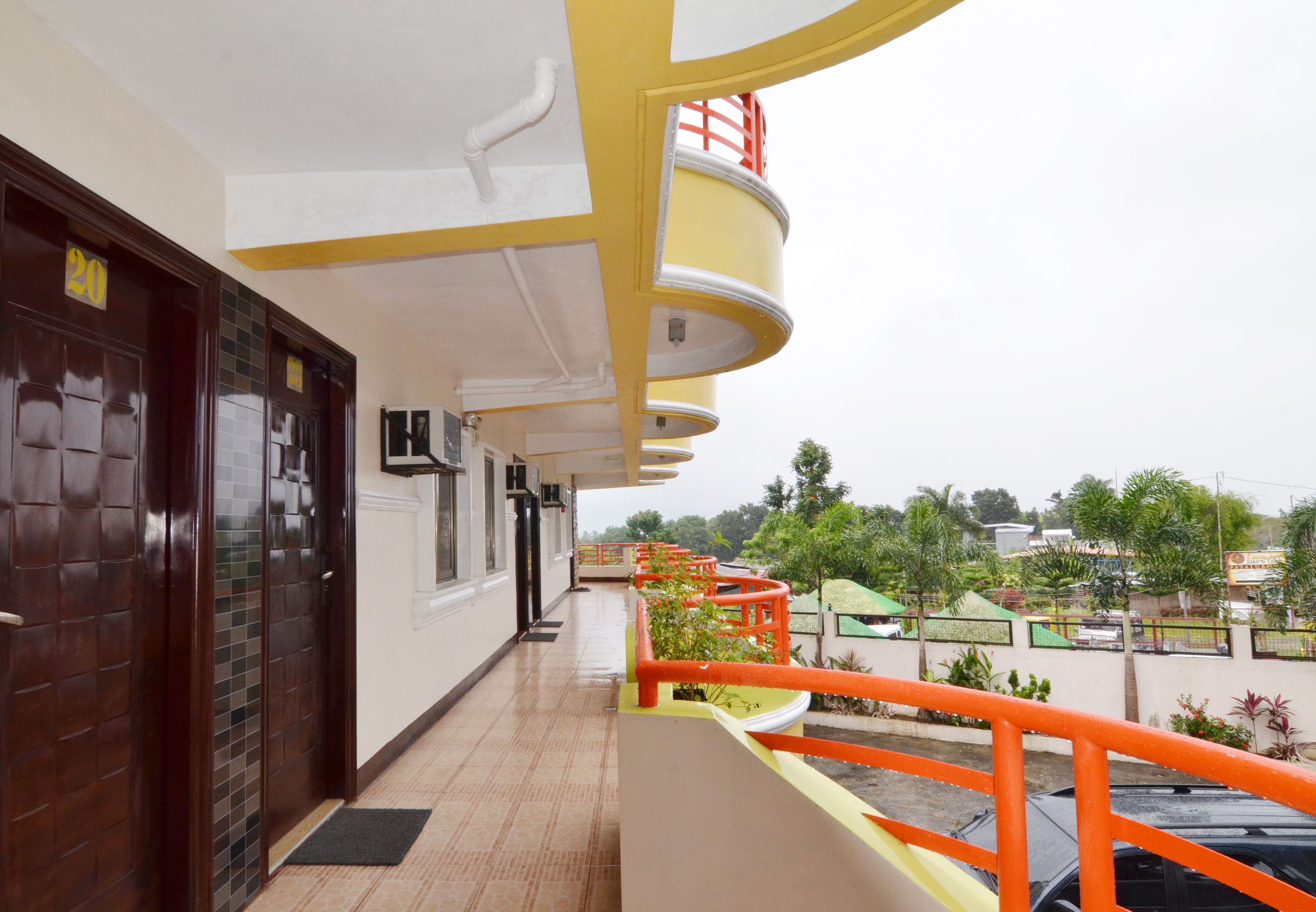 A hotel corridor with a tiled floor, a yellow and white exterior, and a red railing overlooking a green area with trees and a building in the background.