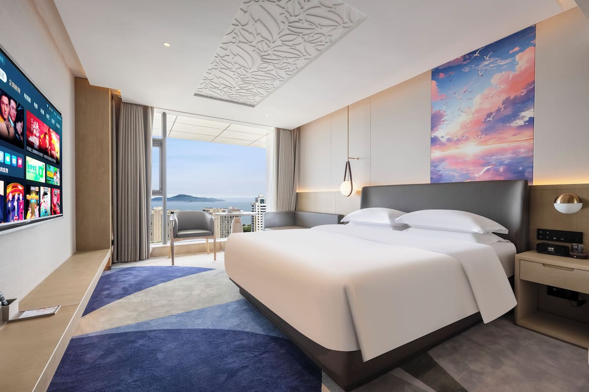 A modern hotel room with a large bed, a TV mounted on the wall, a view of the city, and a large window.