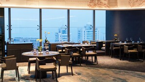 Un espace repas avec des tables et des chaises rondes, de grandes fenêtres offrant une vue sur la ville et un sol recouvert de moquette.