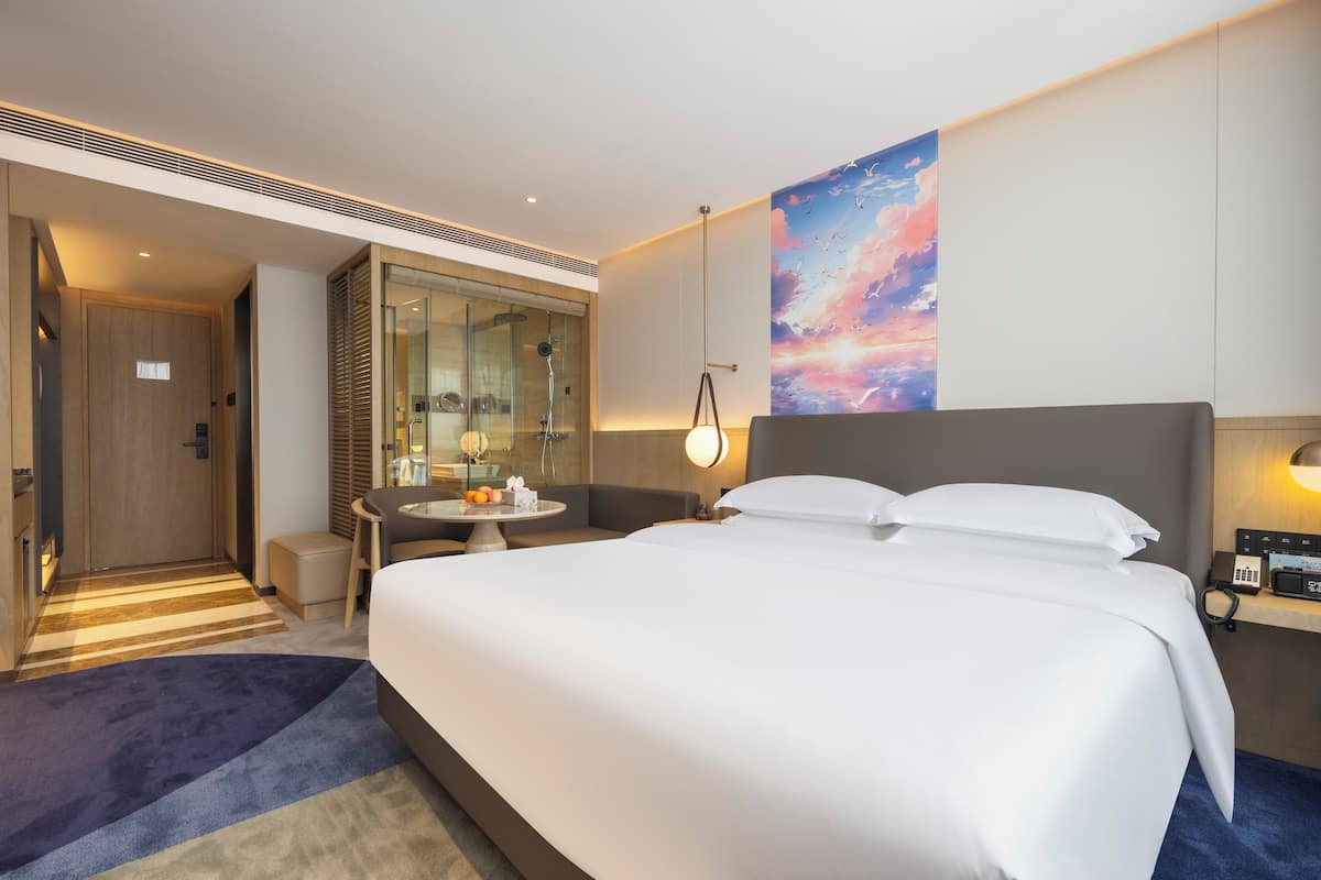 A modern hotel room with a large bed, a small table with a lamp, a shower area, and a painting on the wall.
