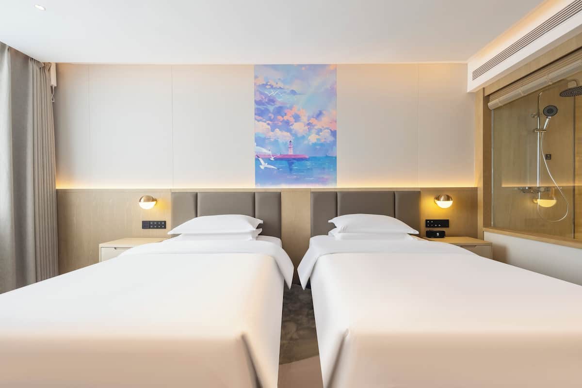 A hotel room with two beds, a painting of a lighthouse on the wall, and a shower area.
