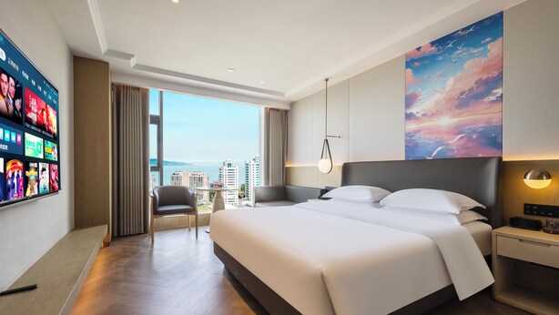 Une chambre d’hôtel moderne, dotée d’un grand lit, d’un téléviseur mural, d’un bureau et offrant une vue sur la ville.