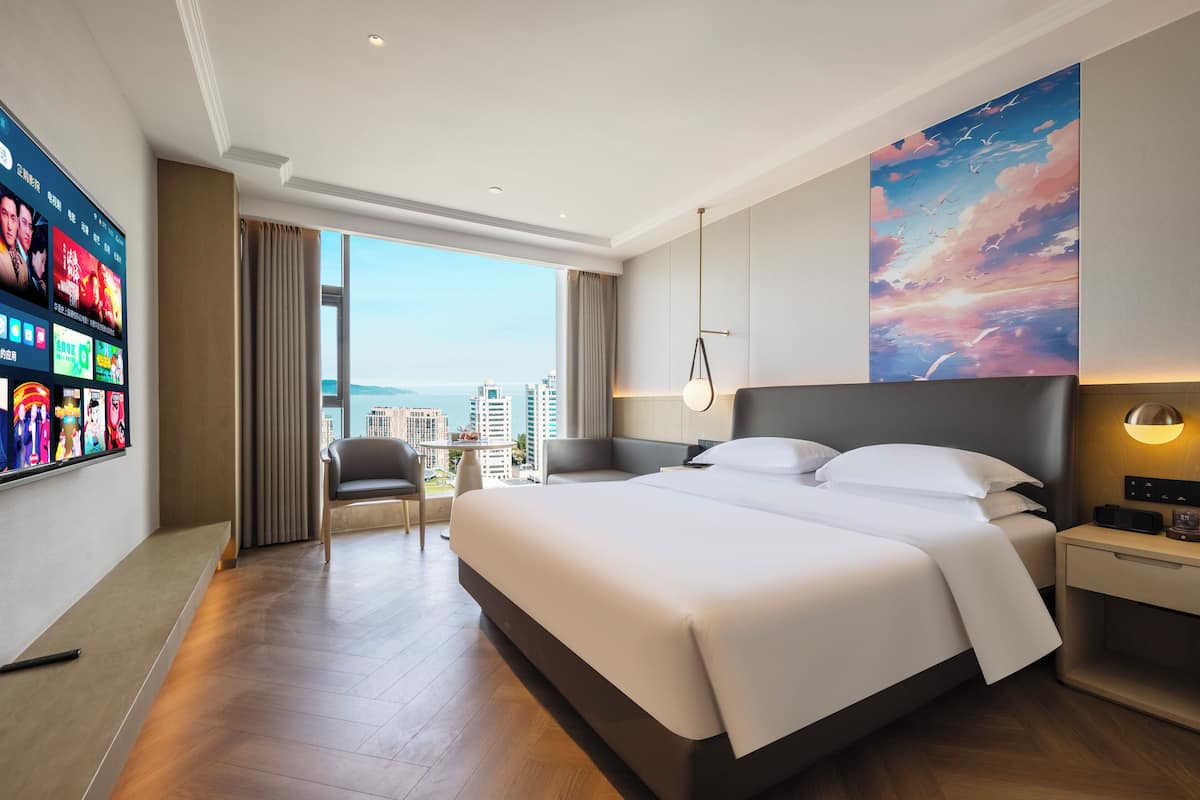 A modern hotel room with a large bed, a TV on the wall, a desk, and a view of the city.