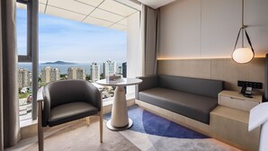 Une chambre d’hôtel moderne dotée d’une grande fenêtre, d’un canapé, d’un fauteuil, d’une petite table et d’une table de chevet avec une lampe.